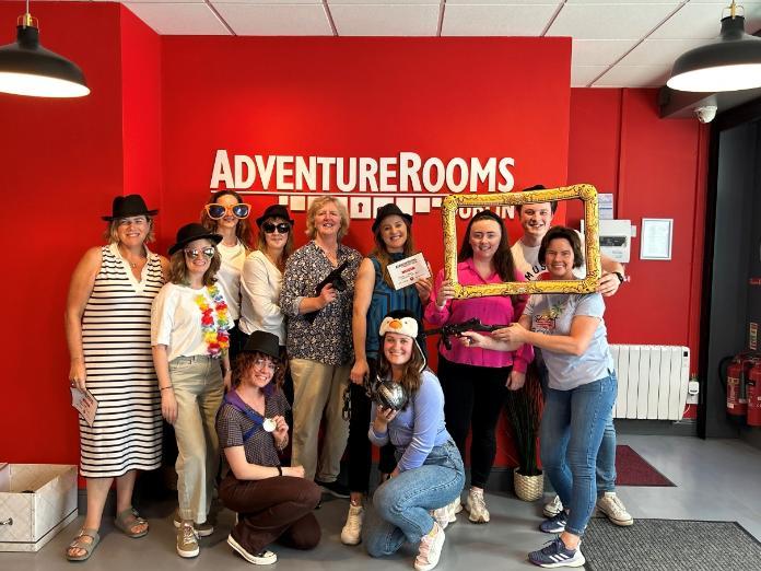 A group photo of the CCRT team at the adventure rooms escape room business. The team are holding mafia related props, which was the theme of the escape room.